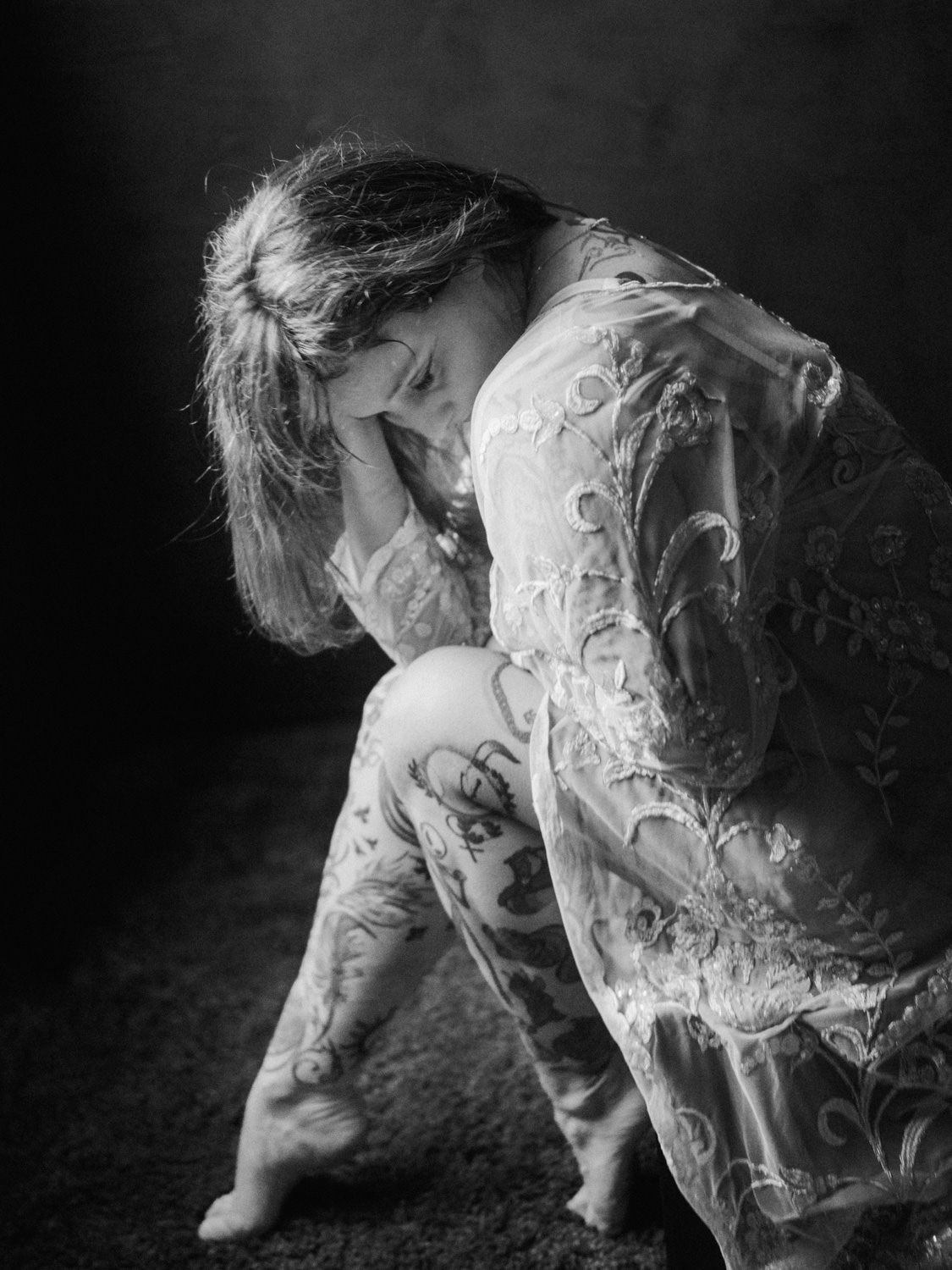 Black and white photo of a woman in lace dress sitting on floor with head in hands in a melancholic pose.