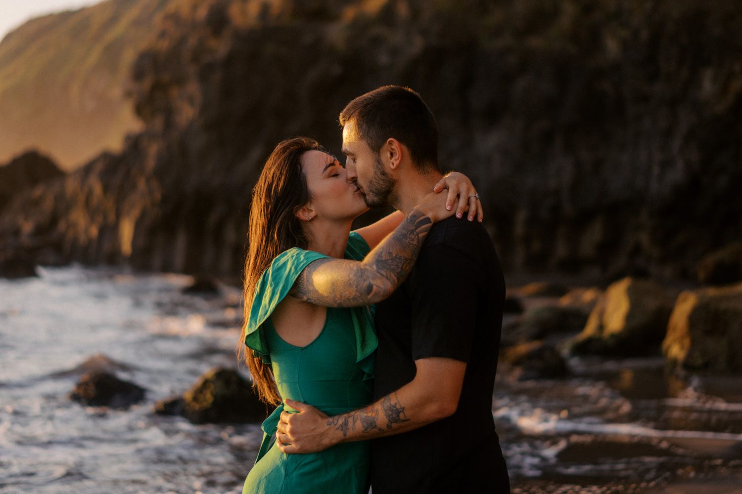 Couple sharing a romantic kiss on a rocky beach at golden hour, woman in green dress with tattoos.