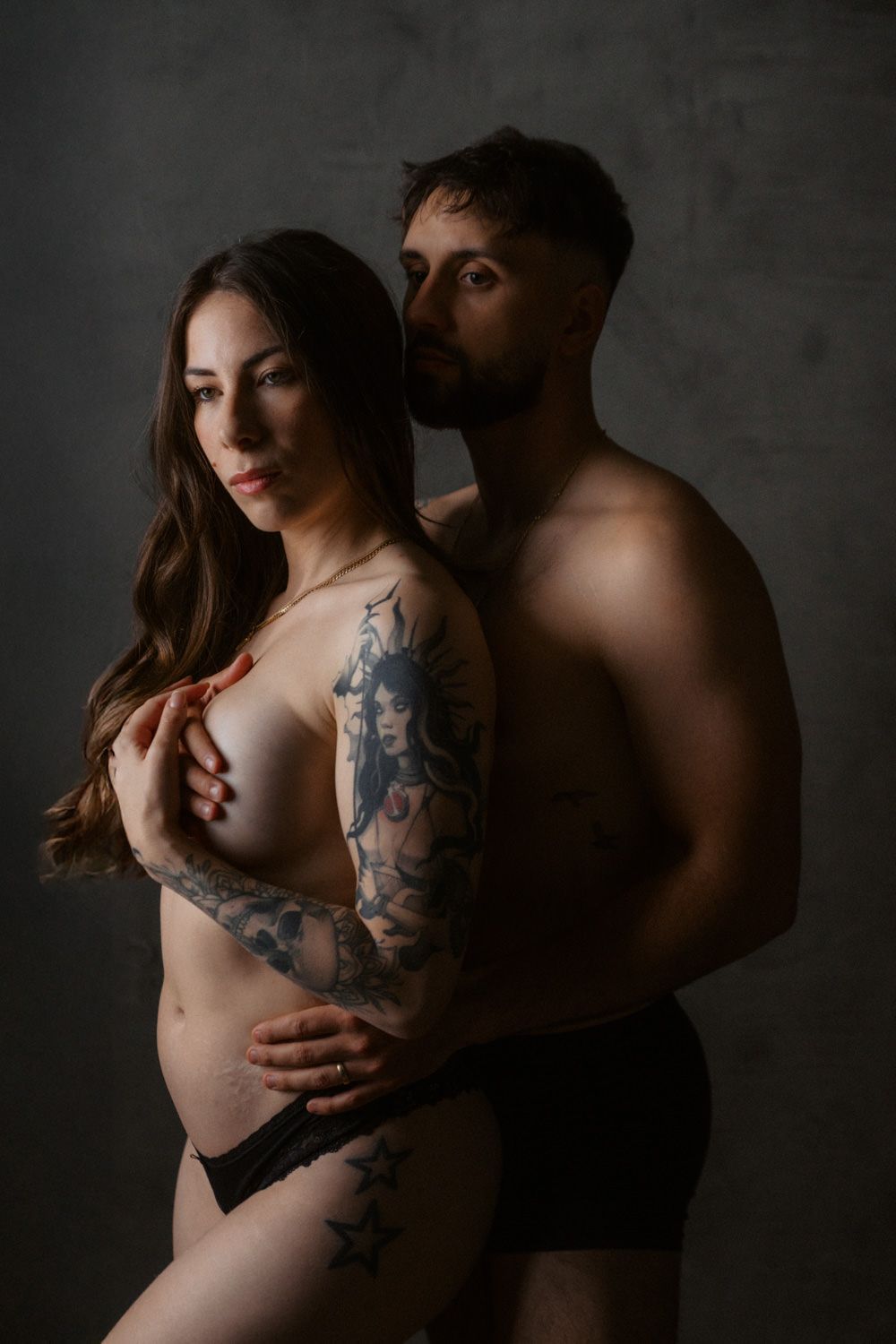 Tattooed pregnant woman poses with shirtless bearded man in intimate maternity portrait against dark background.