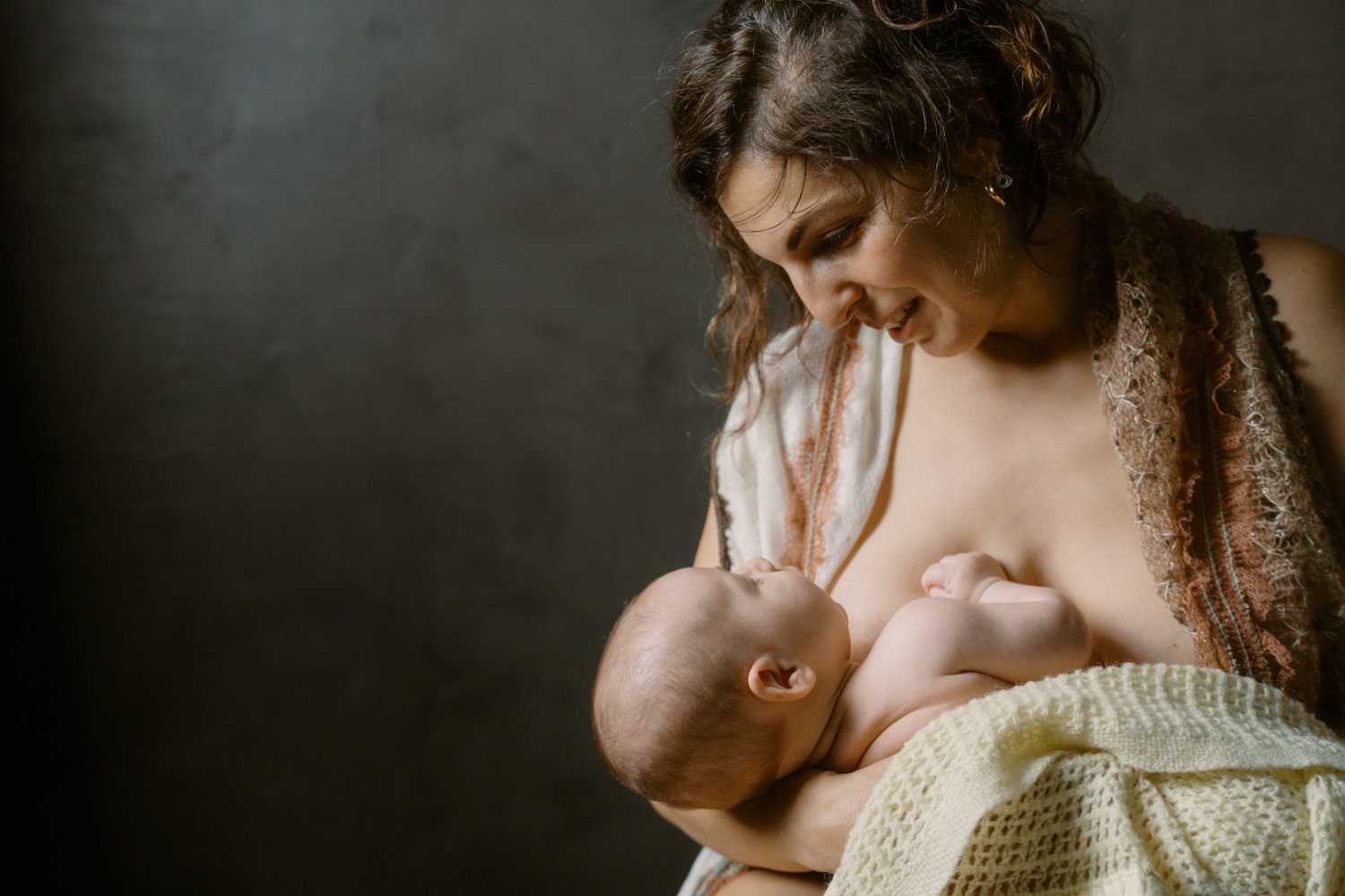Mother tenderly breastfeeding her newborn baby, wrapped in a soft yellow blanket, against a dark background.