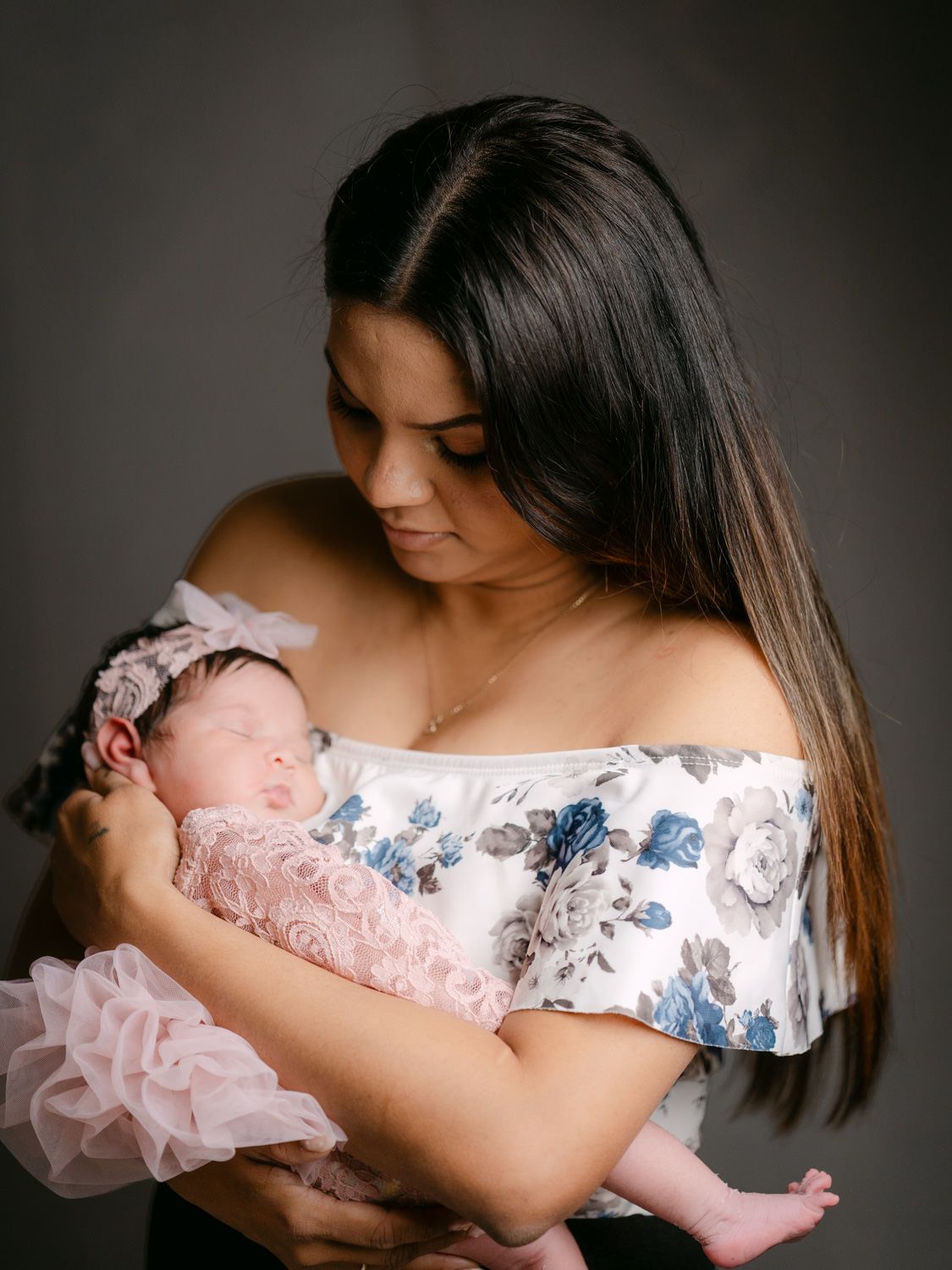 A young mother in a floral off-shoulder top tenderly holds her sleeping newborn dressed in a pink outfit with a bow headband.