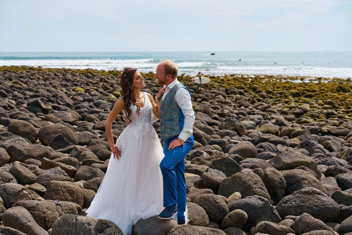 Bride and groom stand on rocky beach shore with ocean waves behind them on their wedding day.