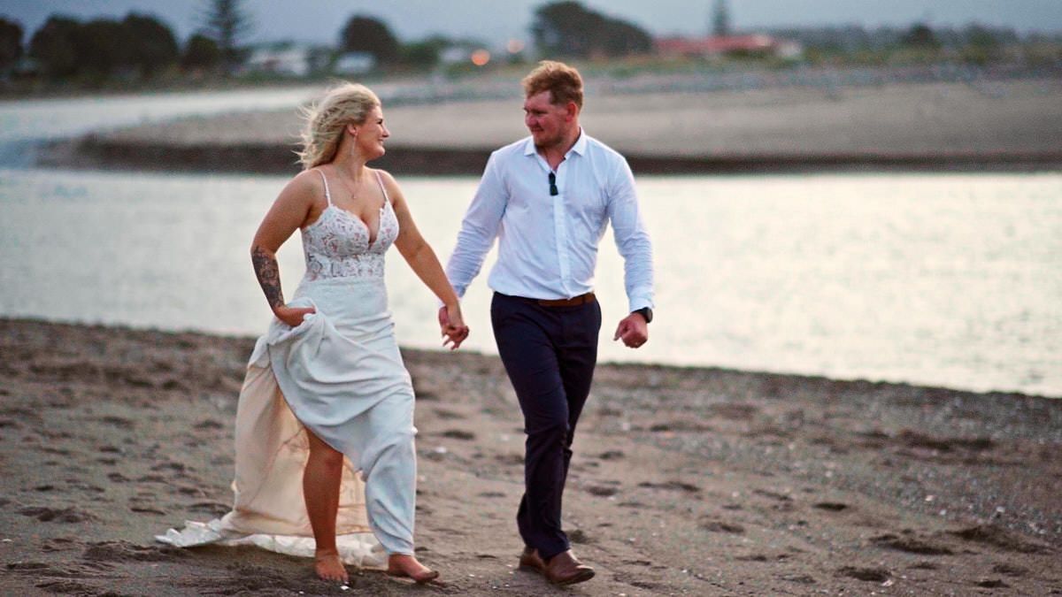 Bride in white dress and groom in white shirt walk hand in hand along a sandy beach at sunset.