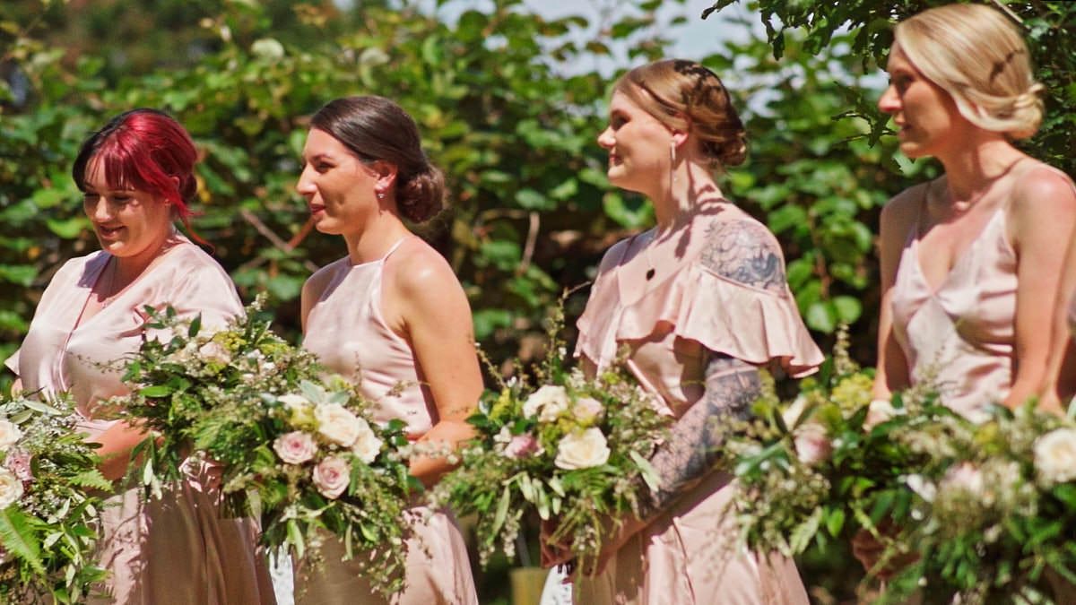 Four bridesmaids in blush pink dresses holding floral bouquets at outdoor wedding ceremony.