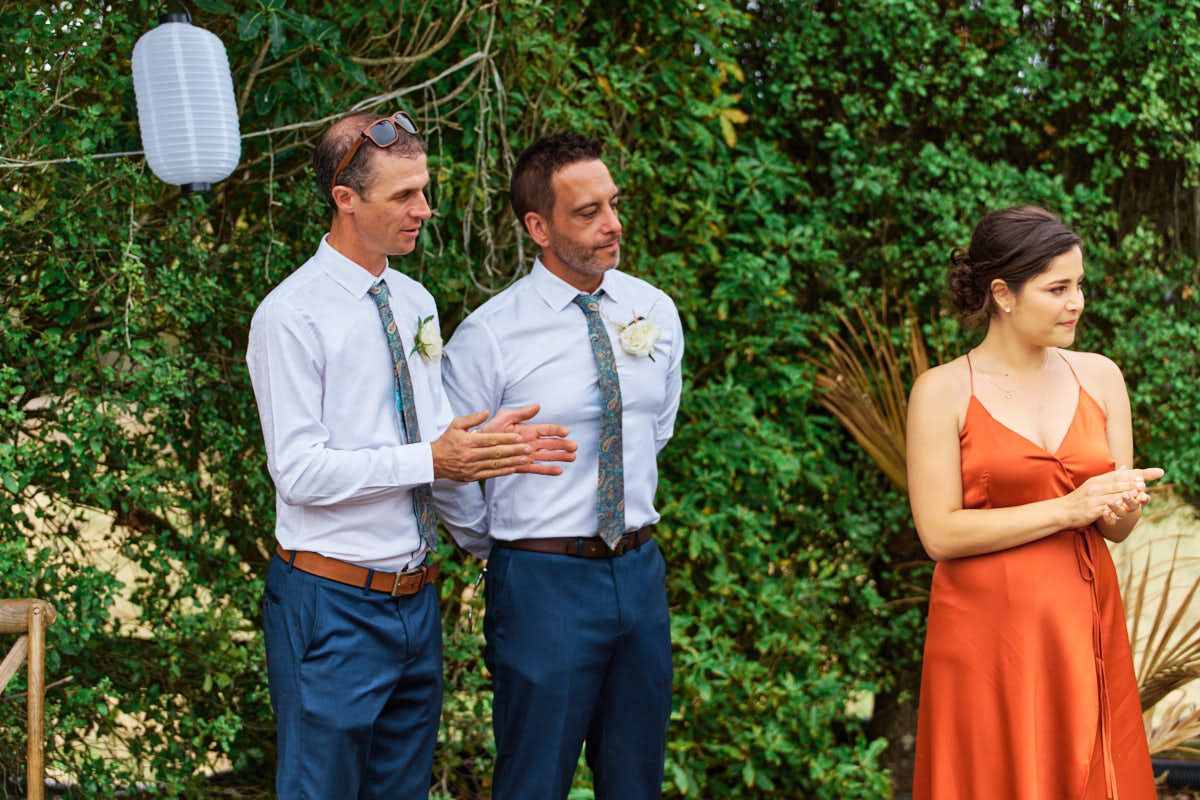 Two grooms in white shirts and navy pants stand with a woman in an orange dress at an outdoor wedding ceremony.