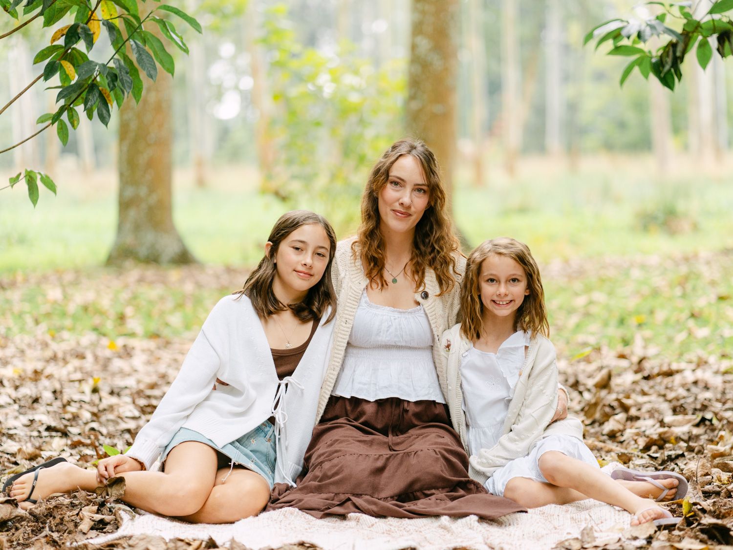 Three girls sitting together on a blanket in a forest, smiling, wearing white and brown outfits.