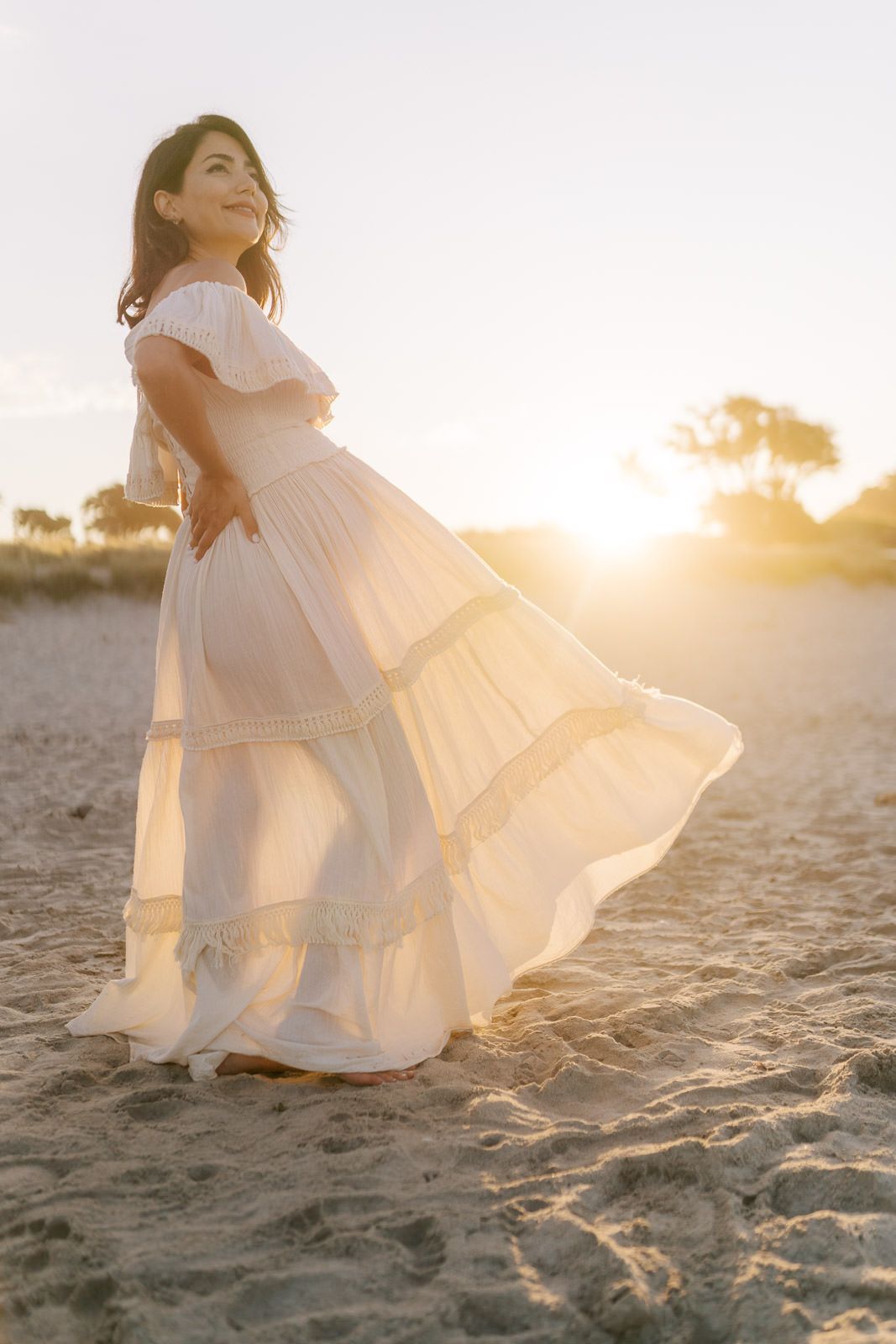 Woman in flowing white dress twirling on sandy beach at golden sunset hour.