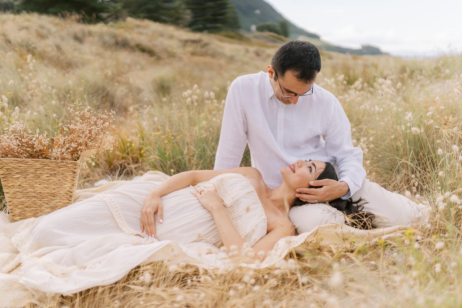 Couple in white attire sharing a romantic moment in a golden meadow, woman lying down smiling up at man.