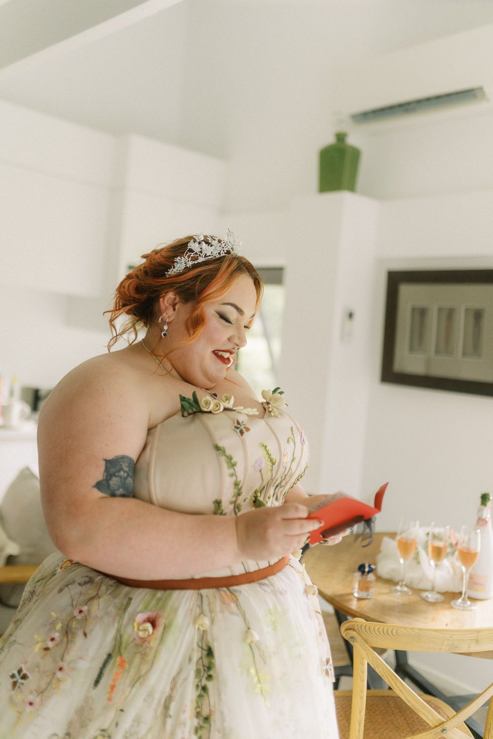 Plus-size bride in floral gown and tiara smiles while reading a card at her wedding reception.