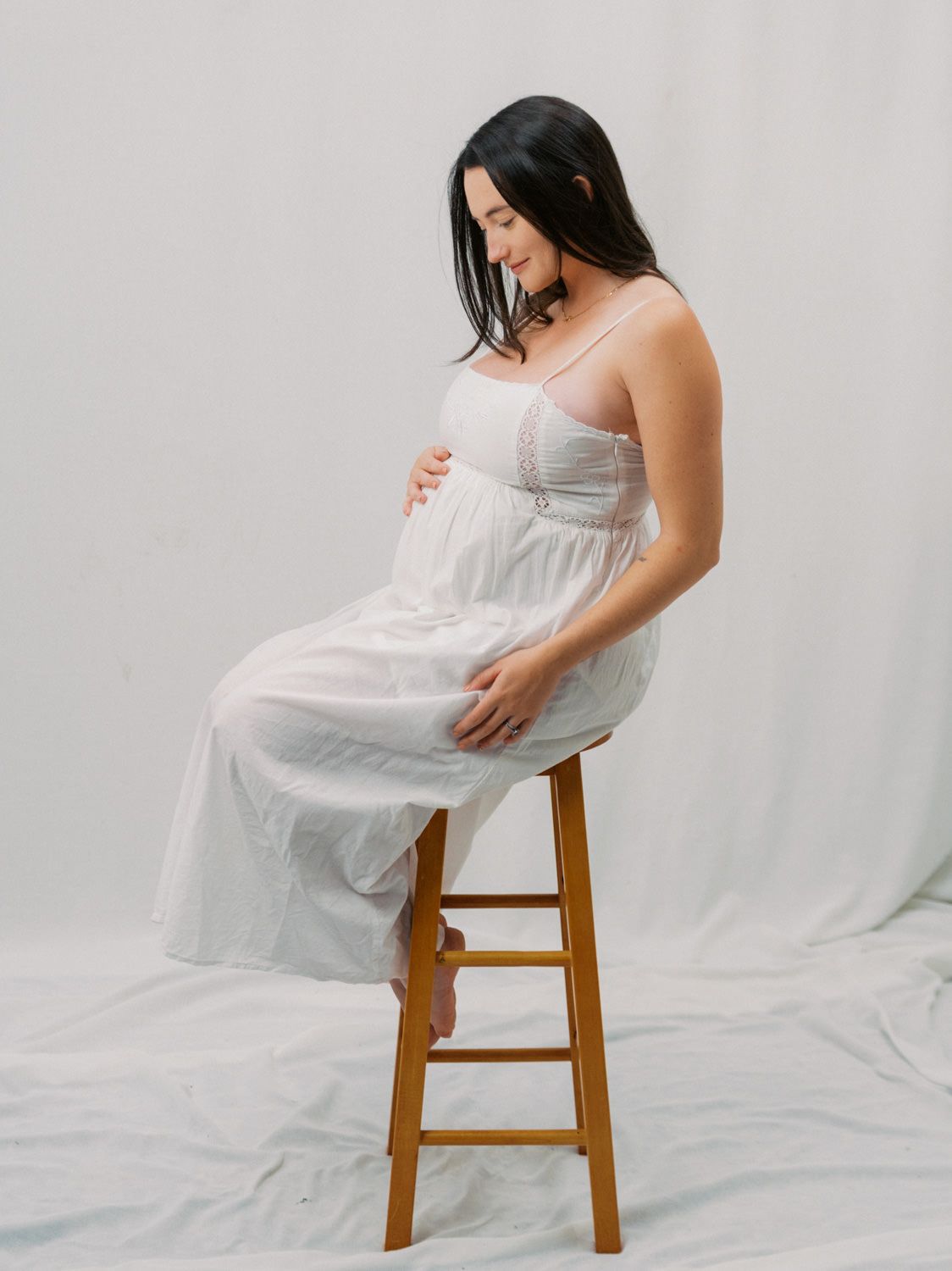 Pregnant woman in white sundress sitting on wooden stool, cradling baby bump during maternity photo shoot.