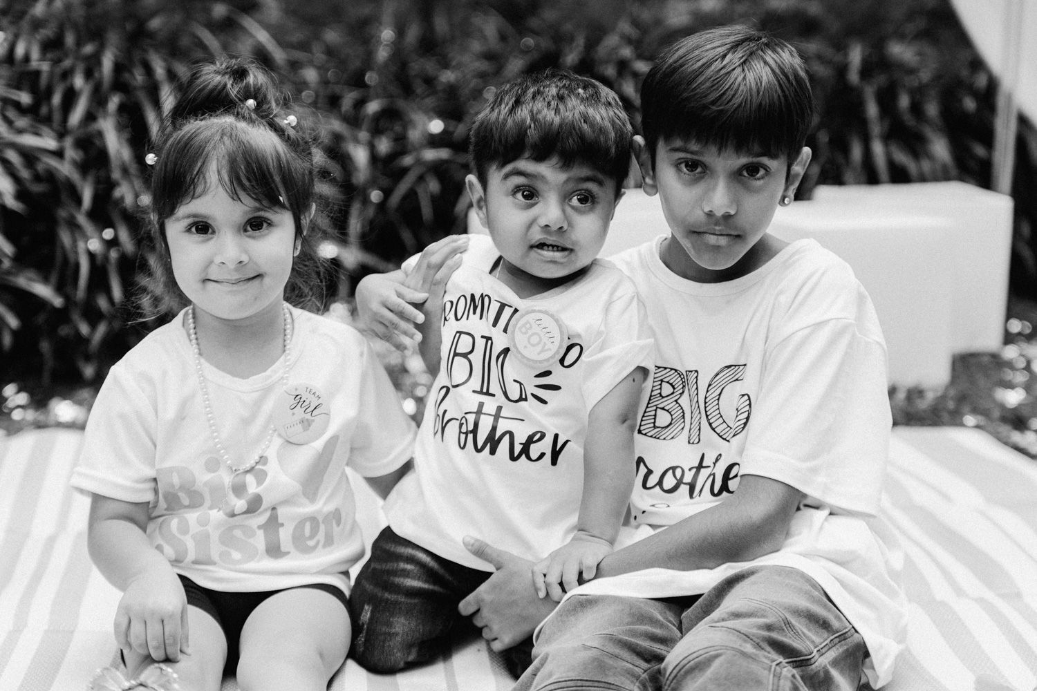 Black and white photo of three young siblings wearing Big Brother and Little Sister shirts, sitting together and smiling.