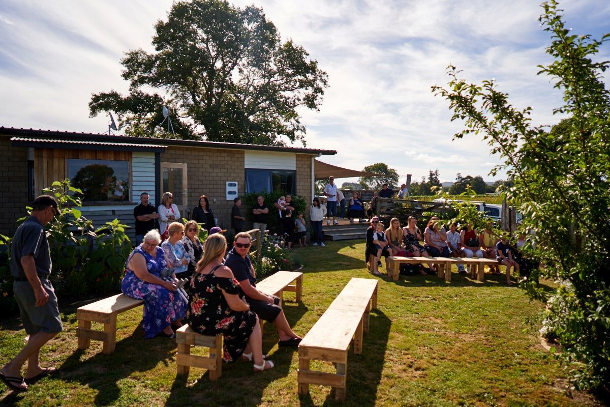 Crowd of people seated on wooden benches outdoors at a sunny community gathering near a wooden building.