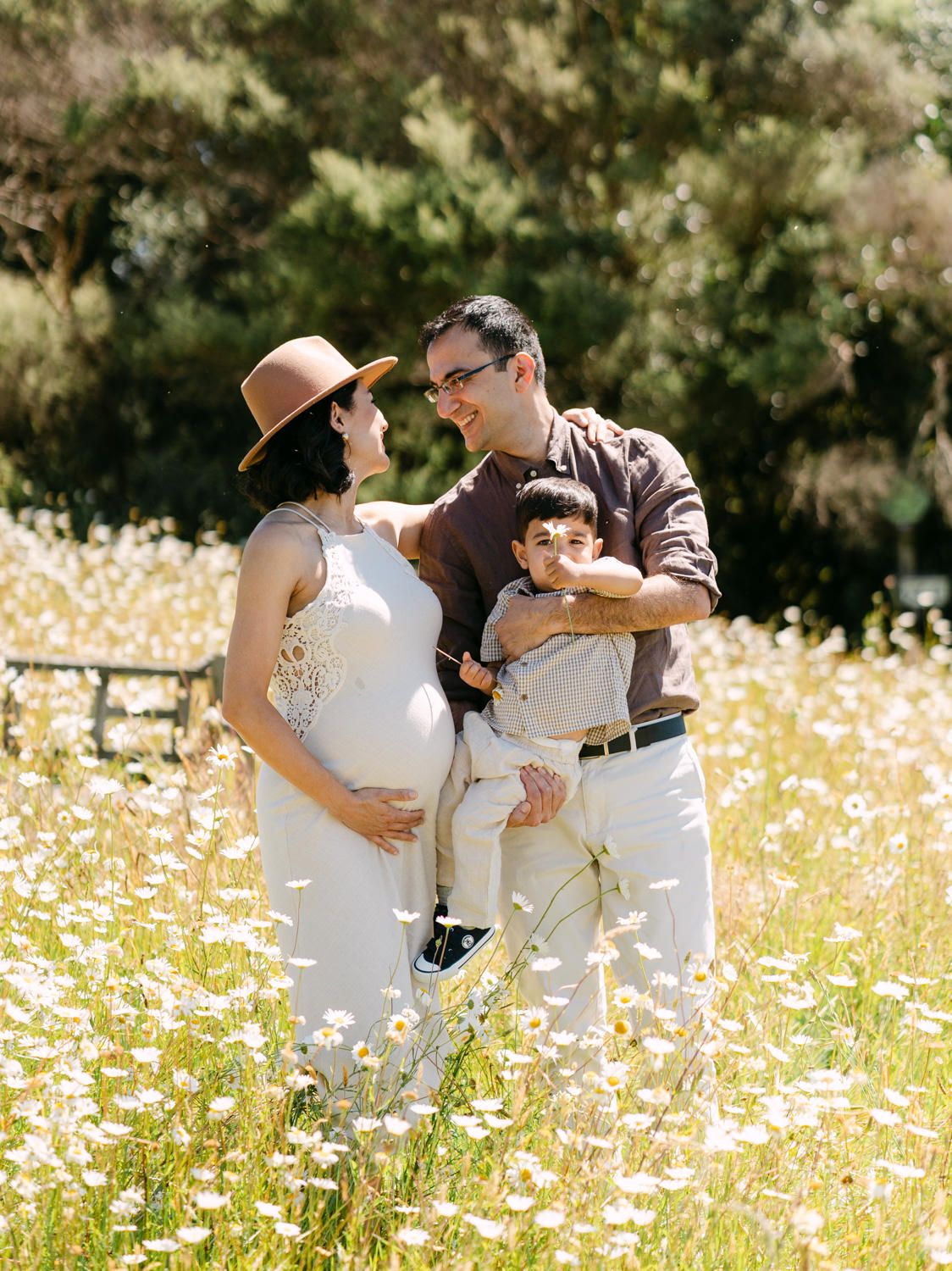Pregnant woman in white dress and hat with husband and toddler son in sunny wildflower field maternity photo.