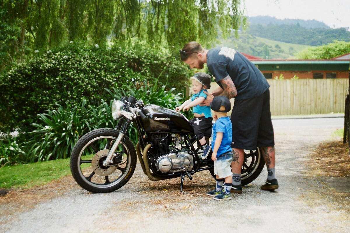 Man showing two young children a black Kawasaki motorcycle on a driveway surrounded by lush green trees.
