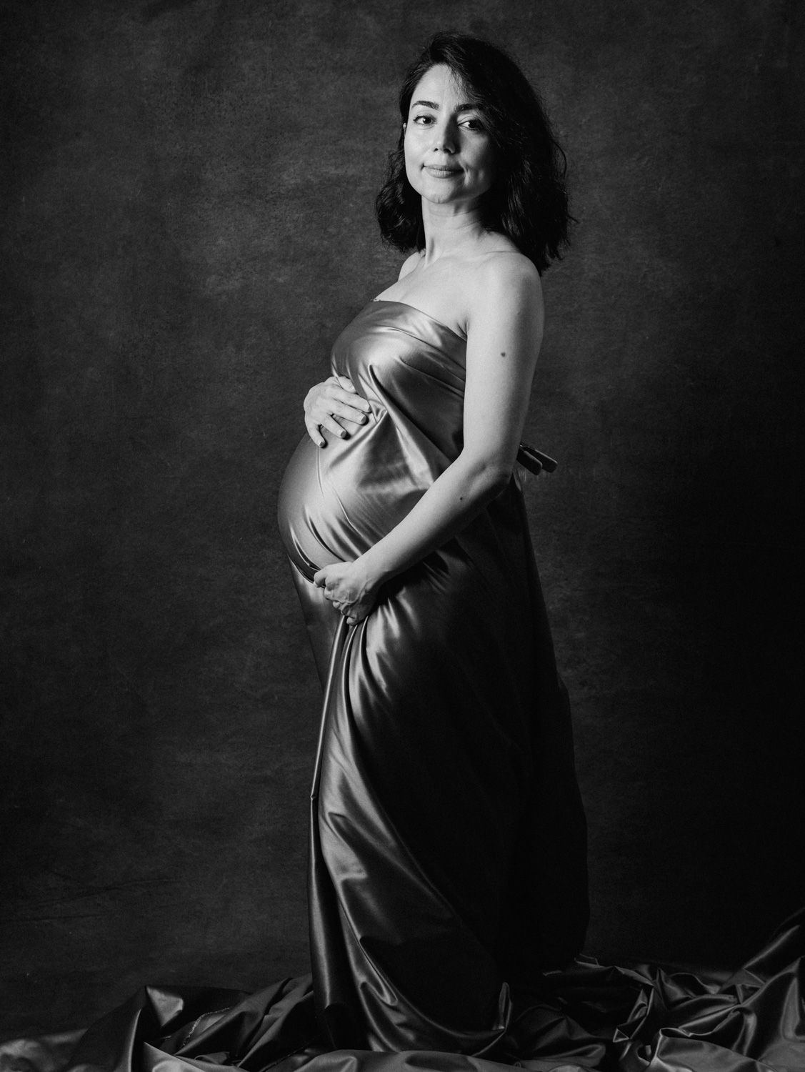 Black and white maternity portrait of a pregnant woman in a satin gown, cradling her baby bump in a studio.
