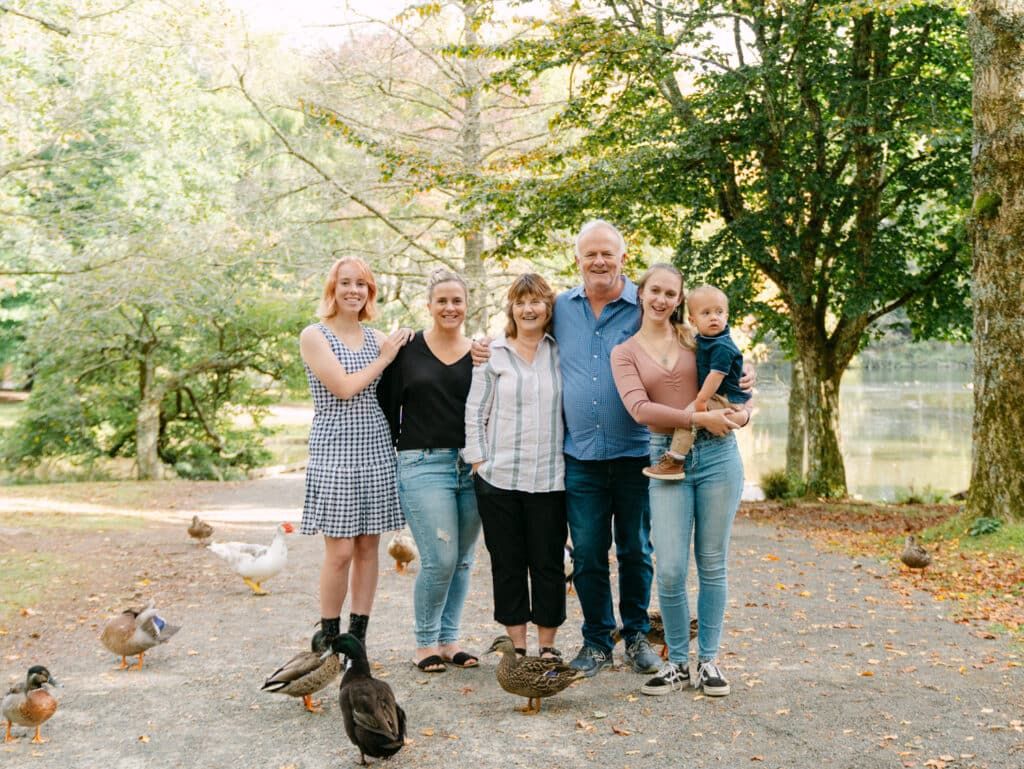 Family of six posing outdoors near a pond surrounded by ducks and trees on a warm day.