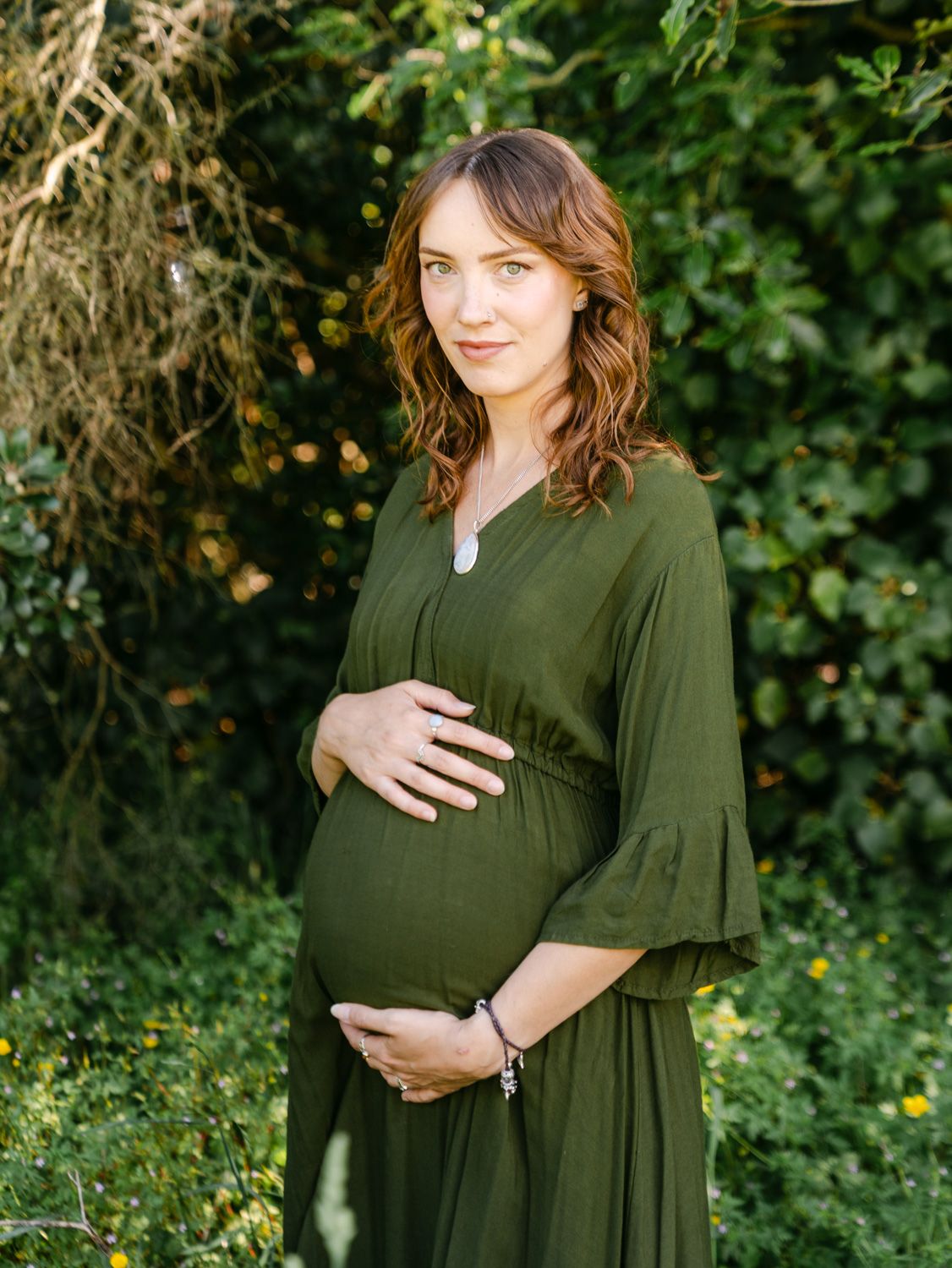 Pregnant woman in olive green dress cradling baby bump outdoors surrounded by lush green foliage.