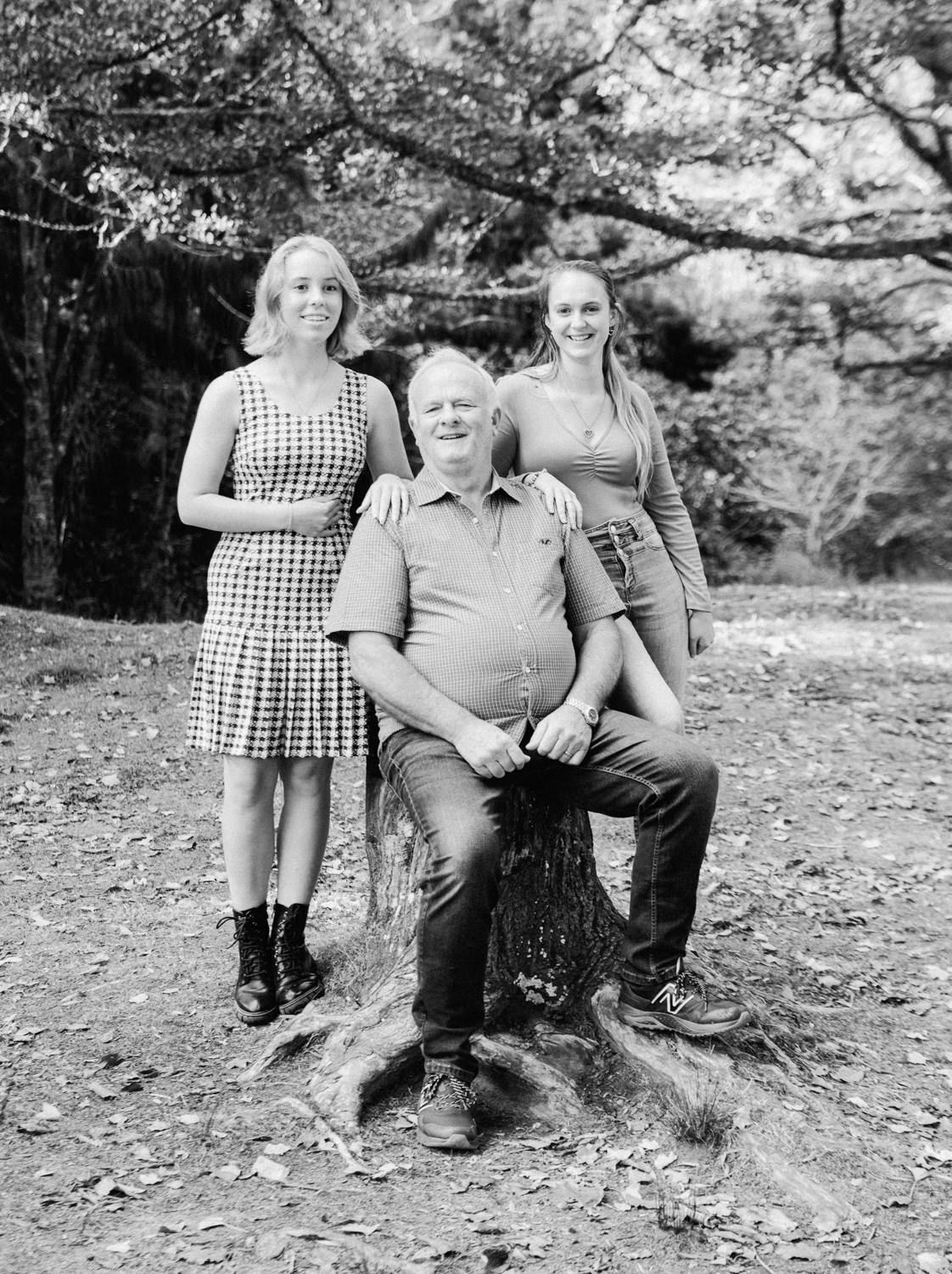 Black and white family portrait of a man seated on a stump with two young women standing beside him outdoors.
