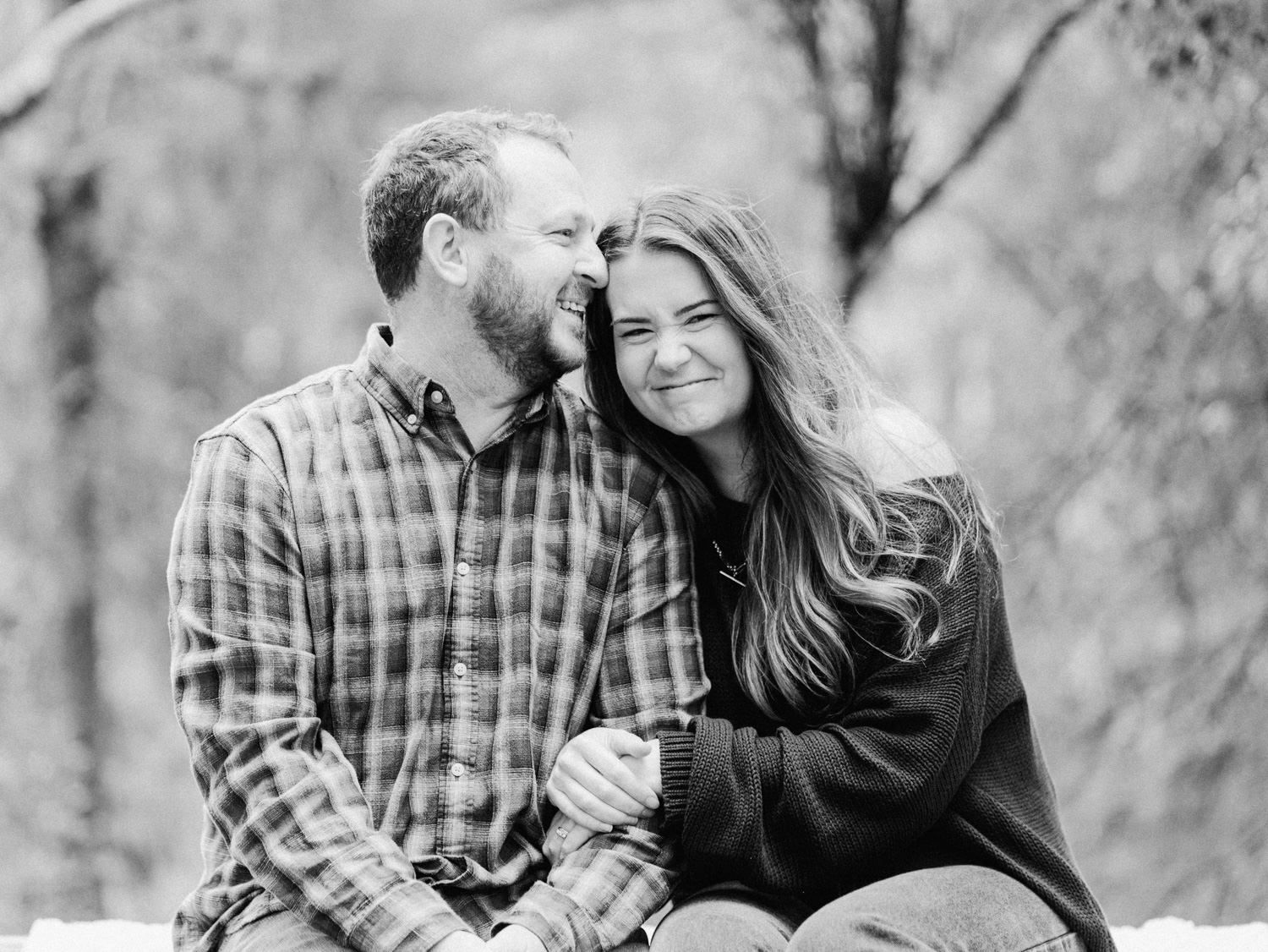 Black and white photo of a smiling couple sitting together outdoors among winter trees.