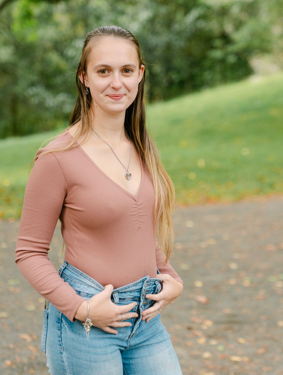 Young woman in pink top and jeans posing outdoors in a park with green trees in background.