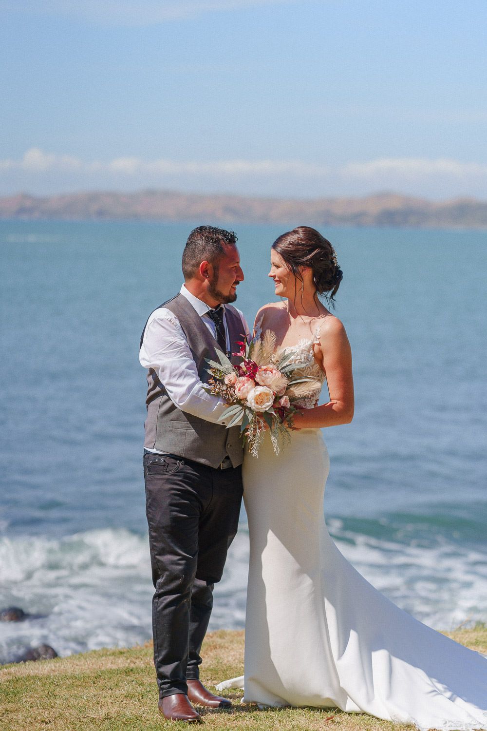 A bride and groom share a romantic moment by the ocean, she holding a floral bouquet in a white gown.