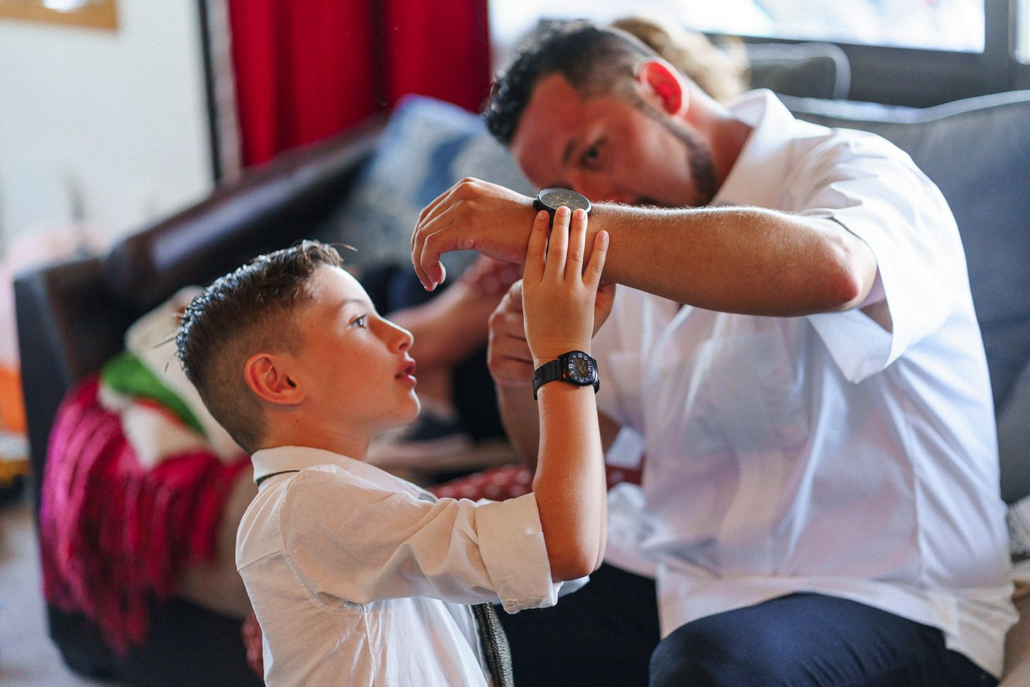 A man helps a young boy adjust his formal white shirt in a bright living room setting.