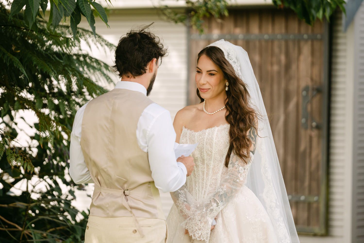 Bride in lace wedding gown and veil exchanging vows with groom in tan vest outdoors near greenery.