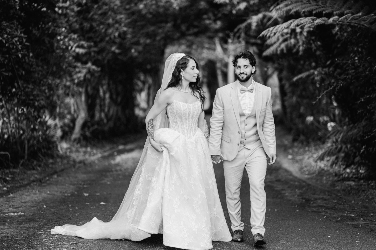 Black and white photo of a bride and groom walking hand in hand down a tree-lined path on their wedding day.