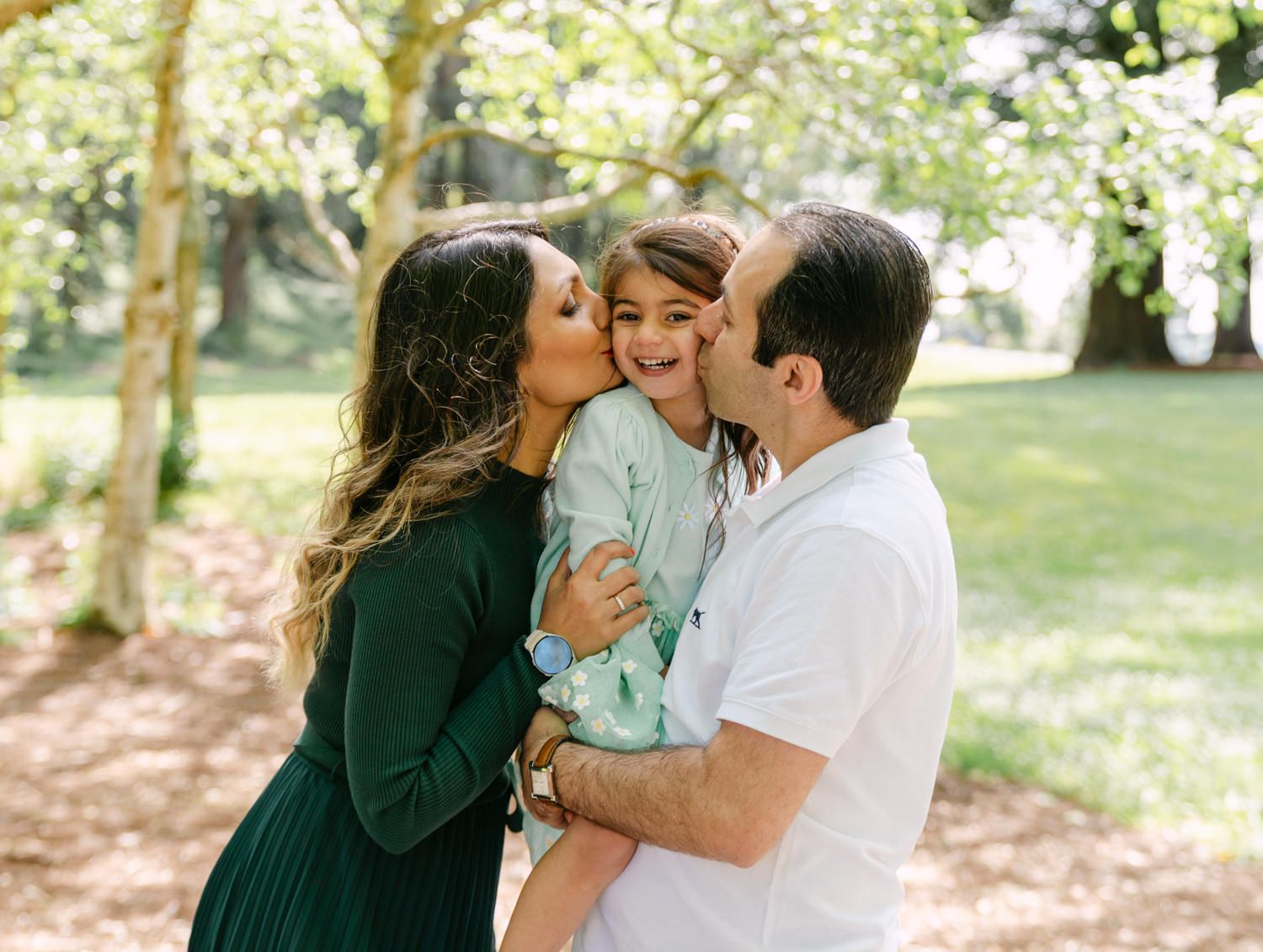 A happy family of three sharing kisses outdoors, with parents kissing their smiling child in a lush green park.