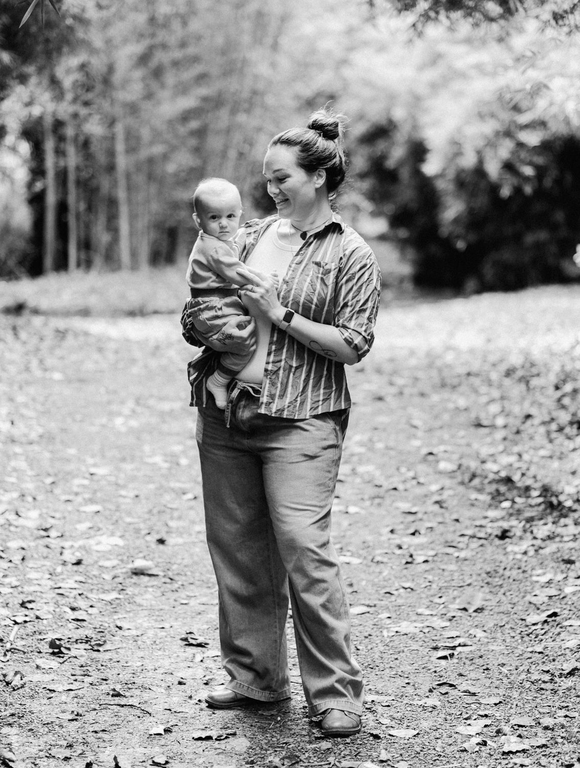 Black and white photo of a smiling woman holding a baby on a leafy forest path.