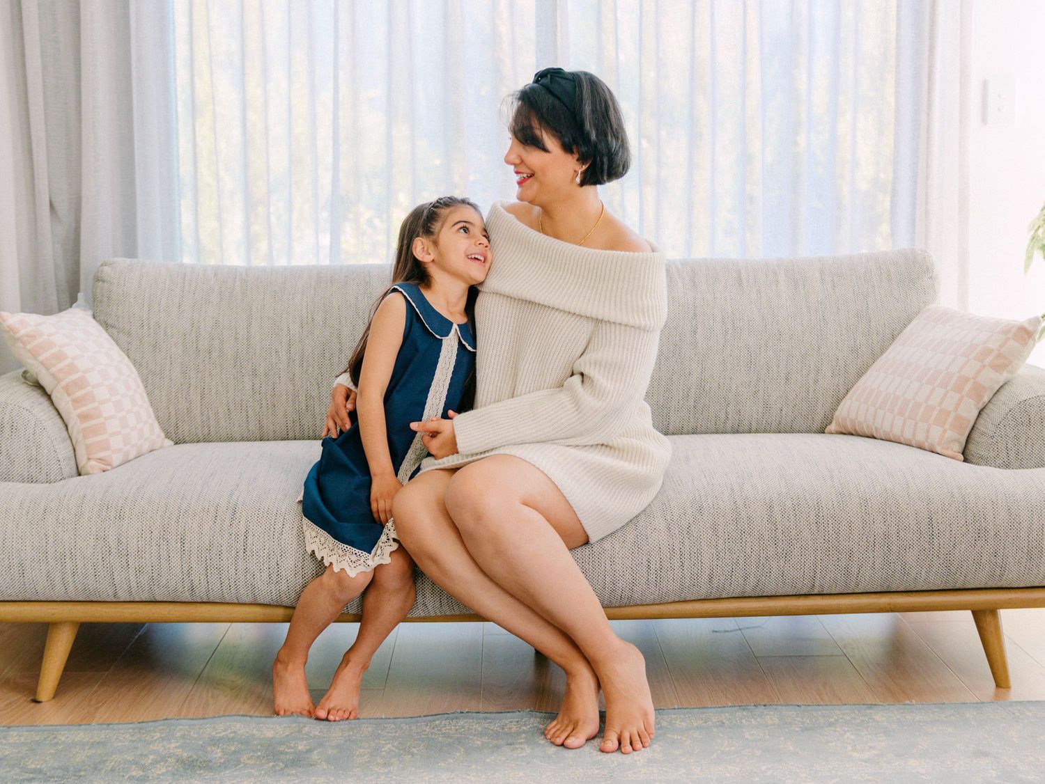 A mother and young daughter share a tender moment sitting together on a modern beige sofa in a bright living room.