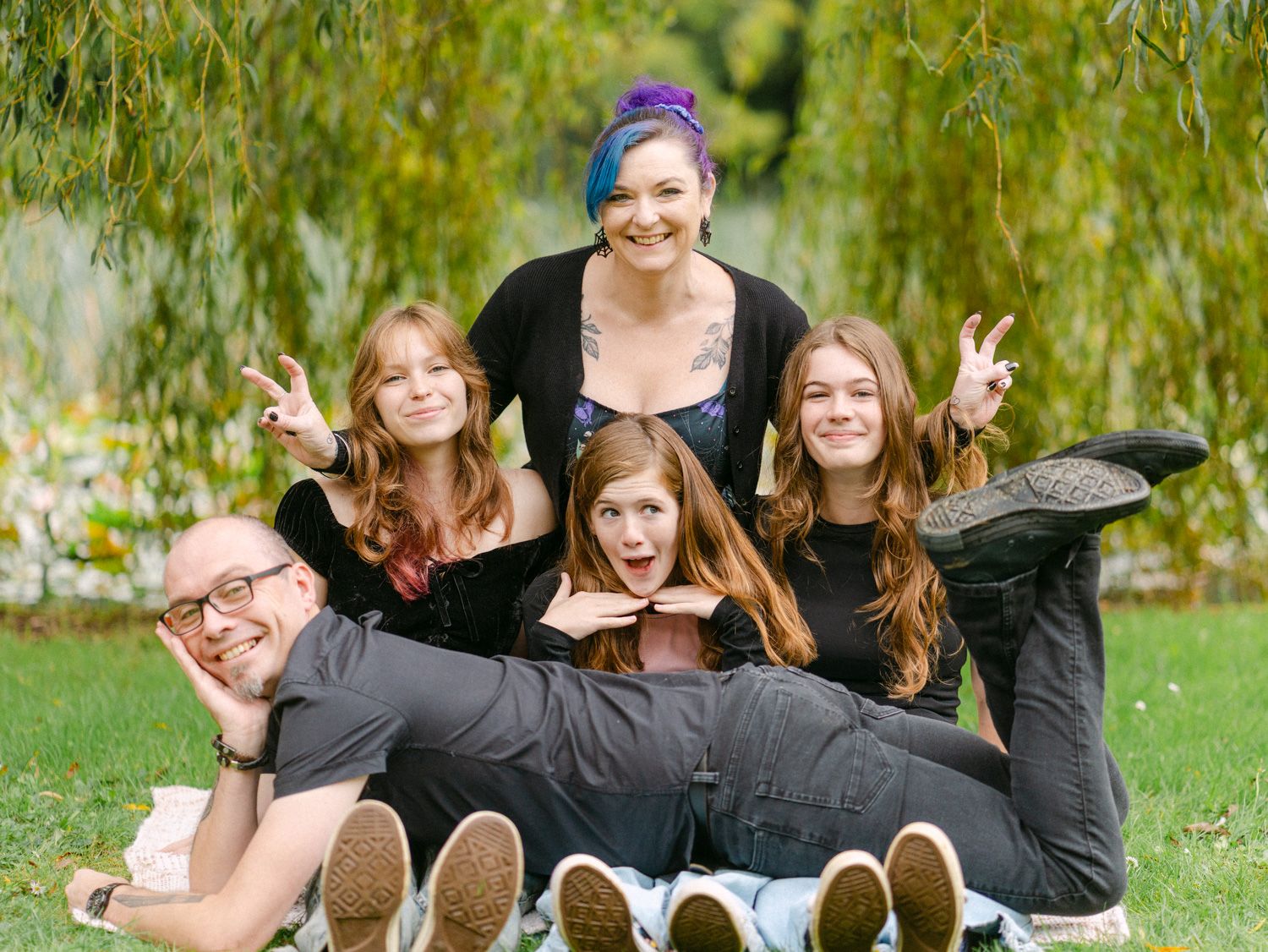 Family of five posing playfully on grass under weeping willow trees, making peace signs in a fun outdoor photo.