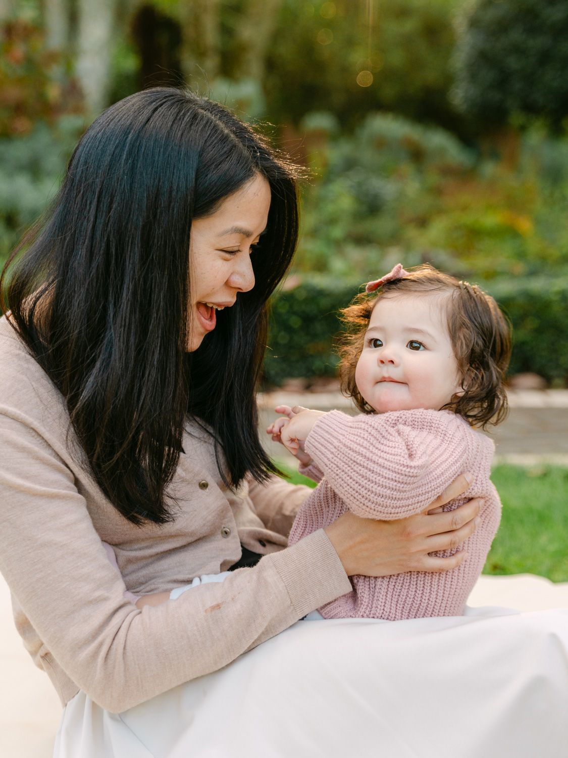 A joyful mother in a pink sweater laughs while holding her adorable baby girl outdoors in a lush garden setting.