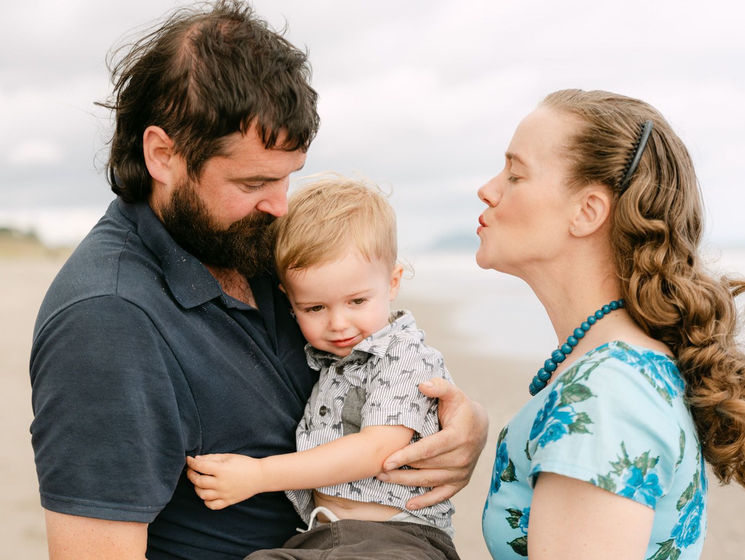 A bearded man and curly-haired woman smile at their toddler boy on a beach.