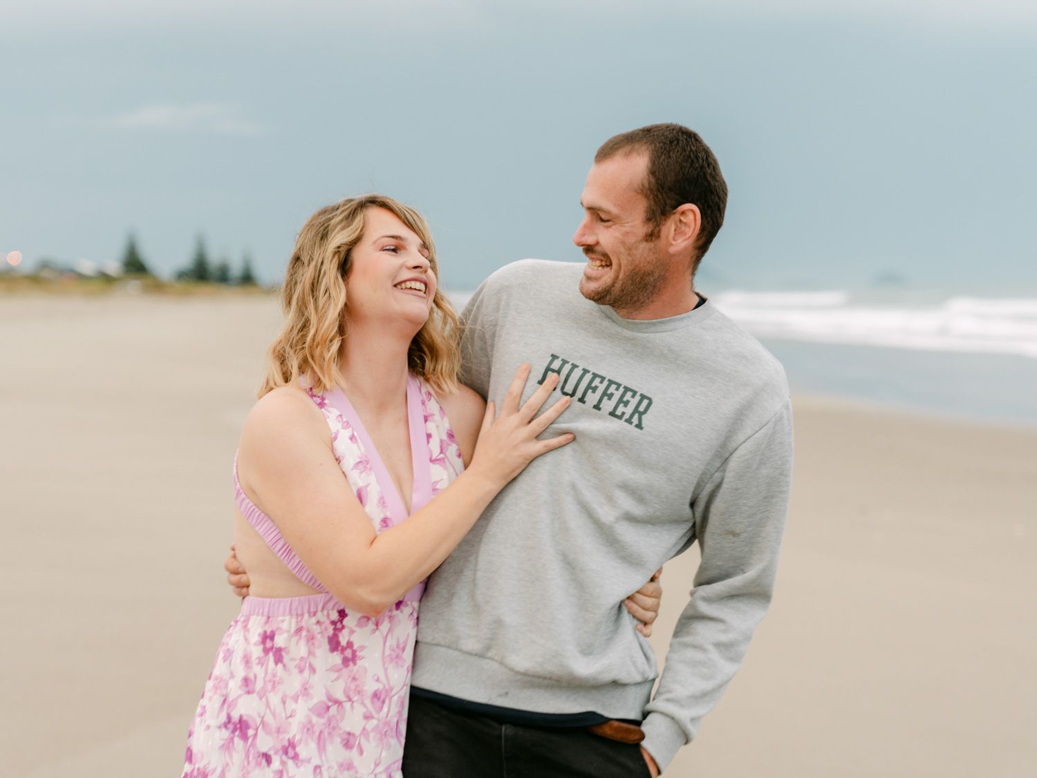 A laughing couple on a beach, woman in pink floral dress and man in grey Huffer sweatshirt, sharing a joyful moment.