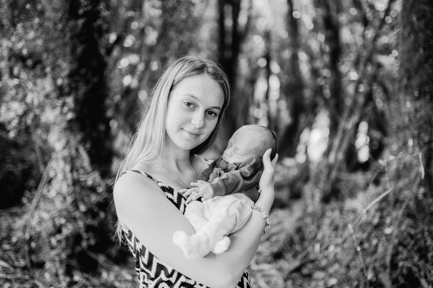 Black and white photo of a young blonde mother tenderly holding her newborn baby in a lush forest setting.