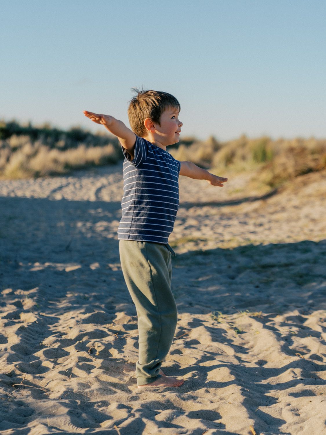 A young boy in a striped shirt and green pants stands on sandy beach with arms outstretched, pretending to fly.