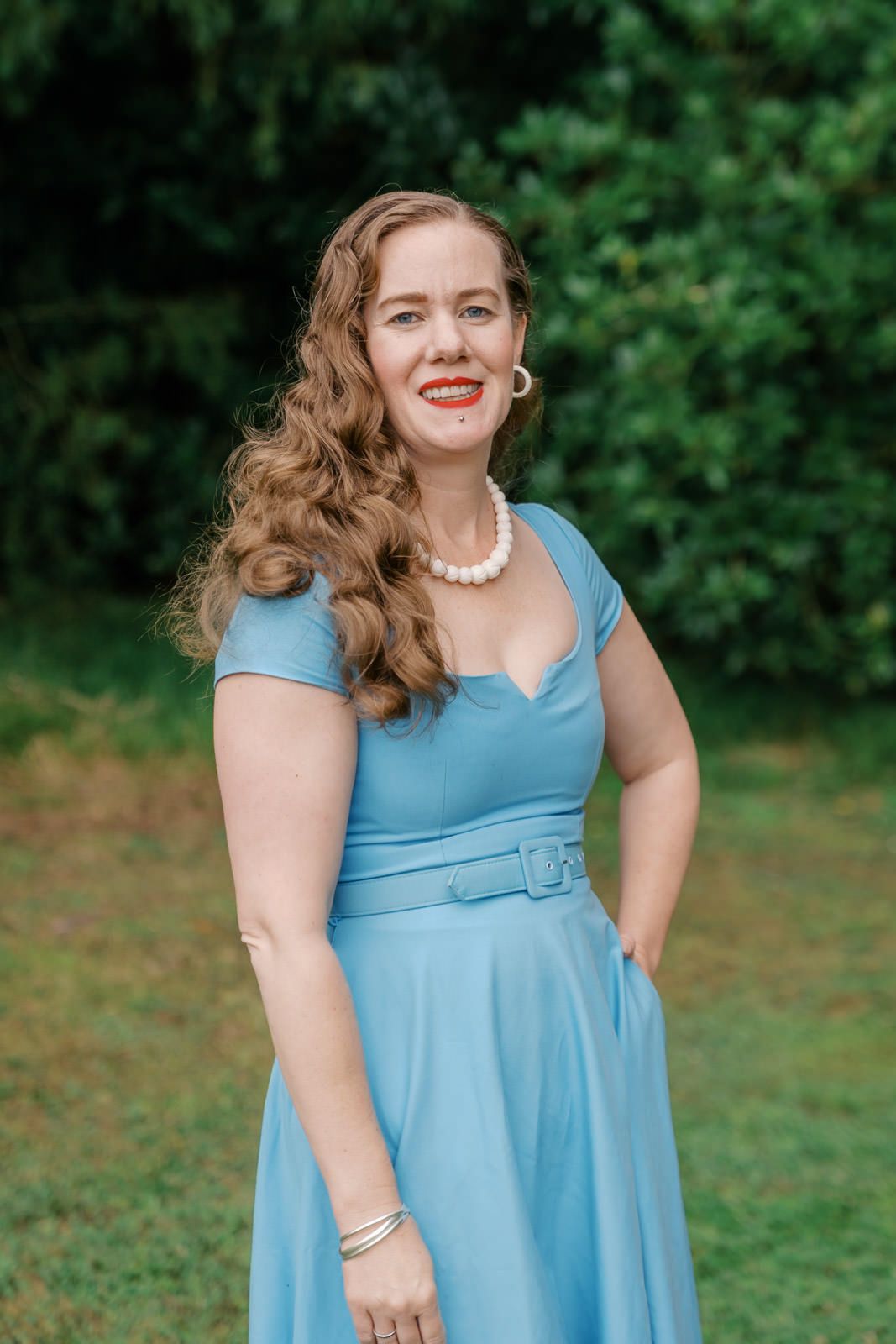 Woman in blue vintage dress with pearl necklace smiling outdoors in garden setting.