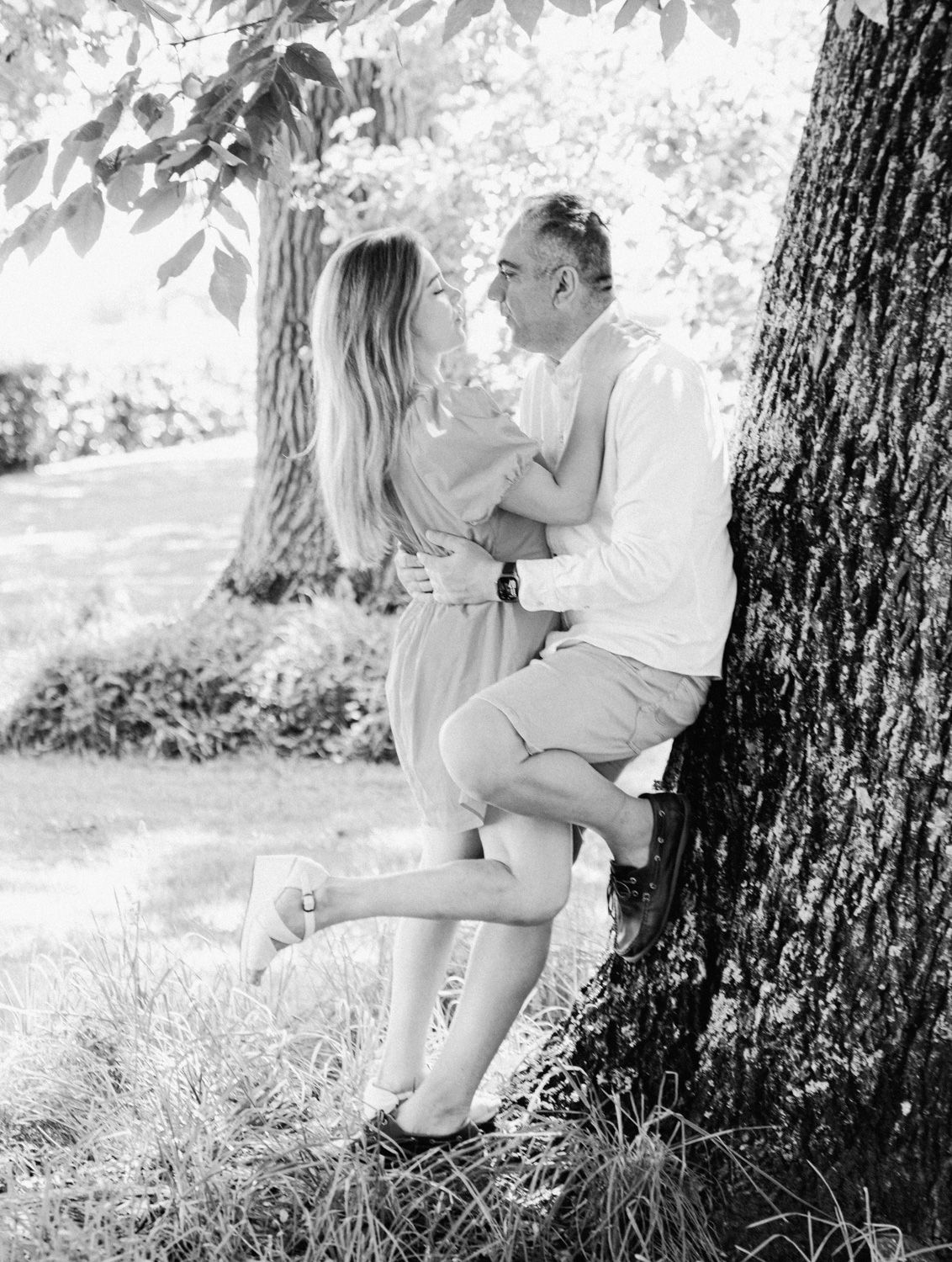 Black and white photo of a couple embracing and laughing together against a large tree in a park.