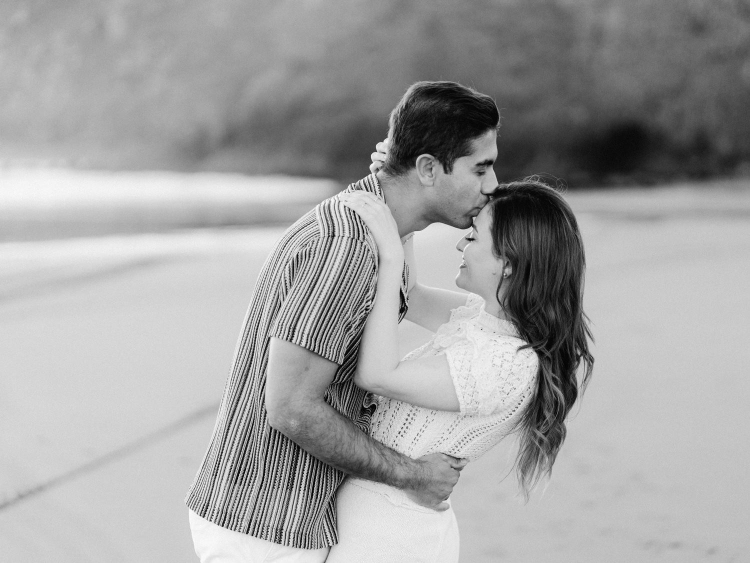 Black and white photo of a romantic couple embracing and kissing on a beach during an engagement session.