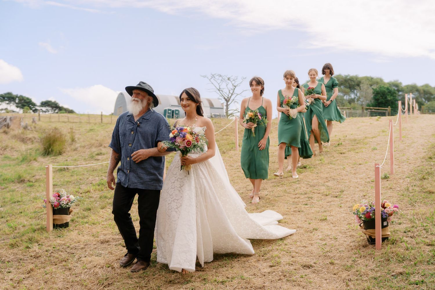 Bride walks down outdoor aisle with older man, bridesmaids in green dresses follow, flower arrangements line the path.