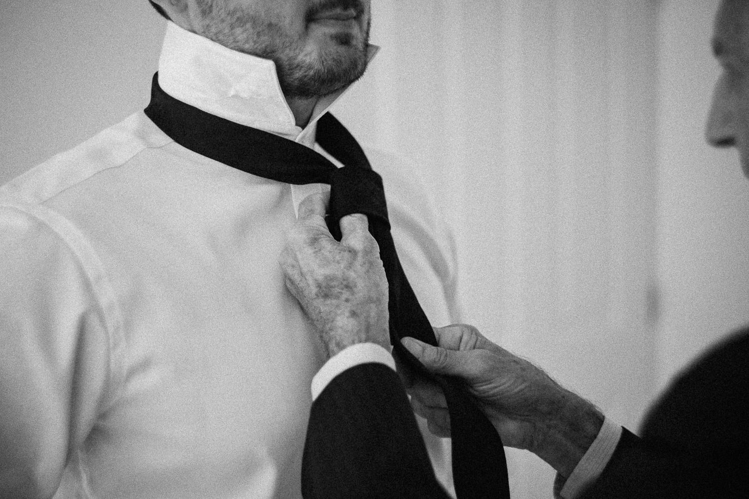 Black and white photo of a man having his tie adjusted by another person, elegant wedding preparation scene.