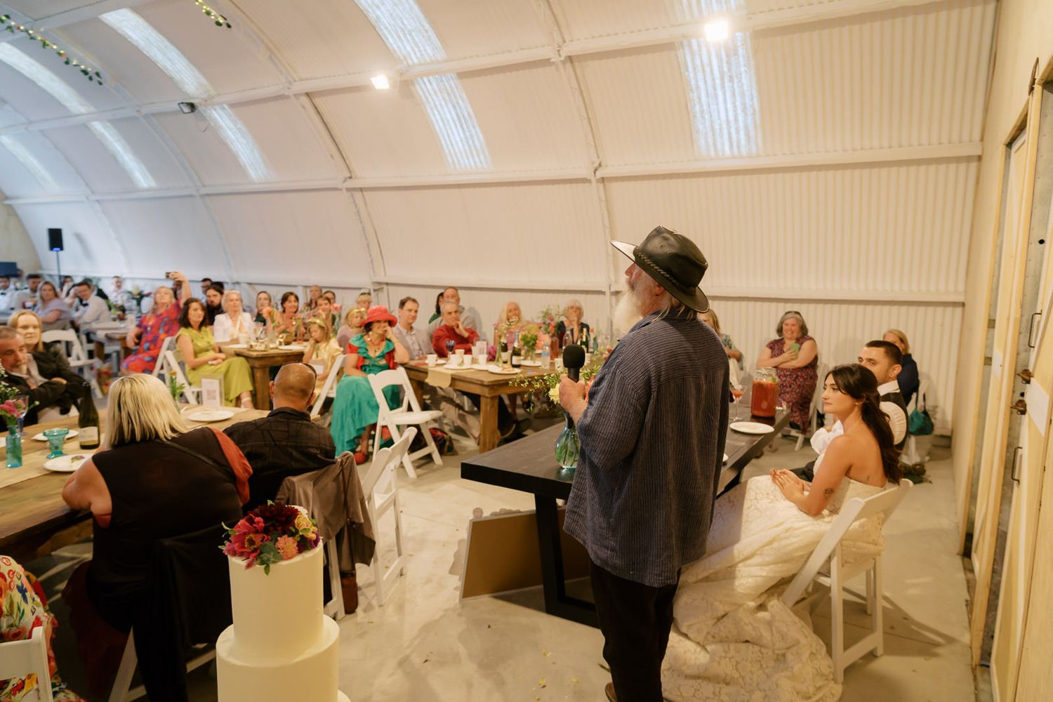 Person speaking at a wedding reception inside a tent venue with guests seated at decorated tables.