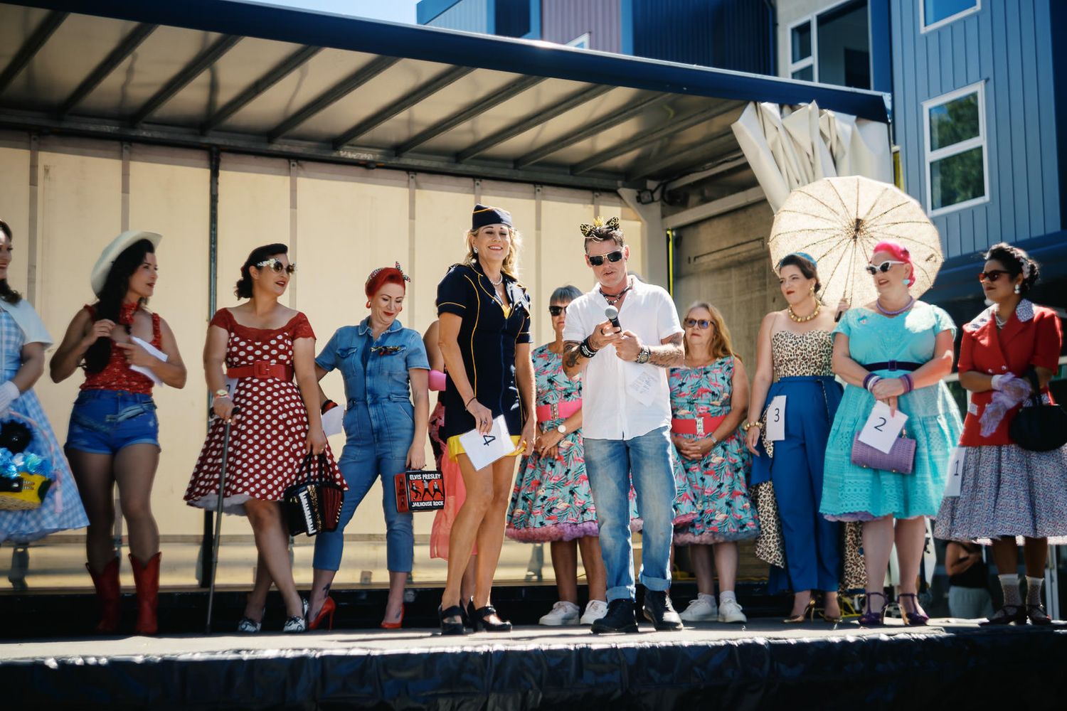 Group of people in 1950s vintage pin-up fashion on stage at a retro-themed outdoor event.