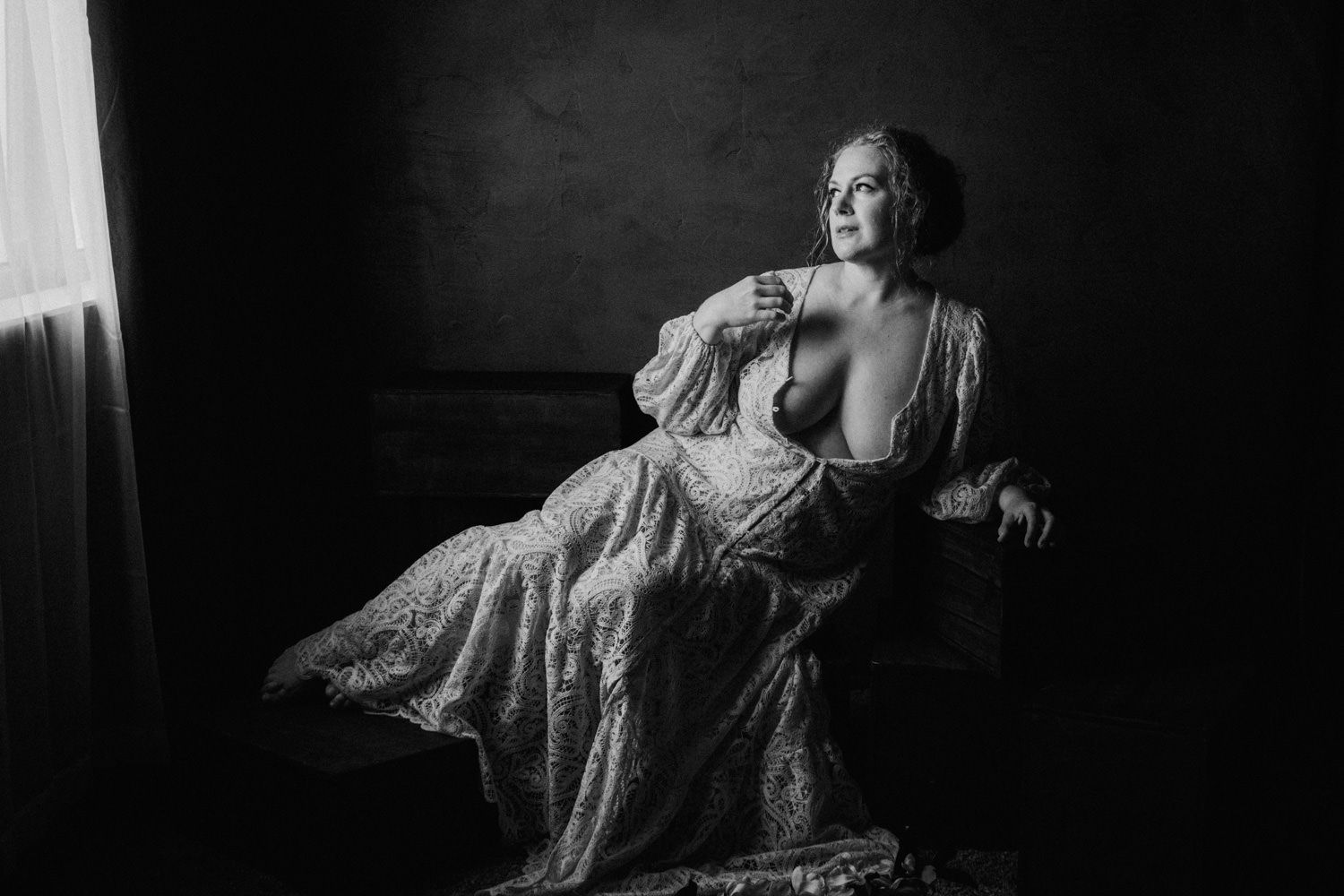 Black and white boudoir portrait of a woman in lace gown seated elegantly by a window in dramatic lighting.
