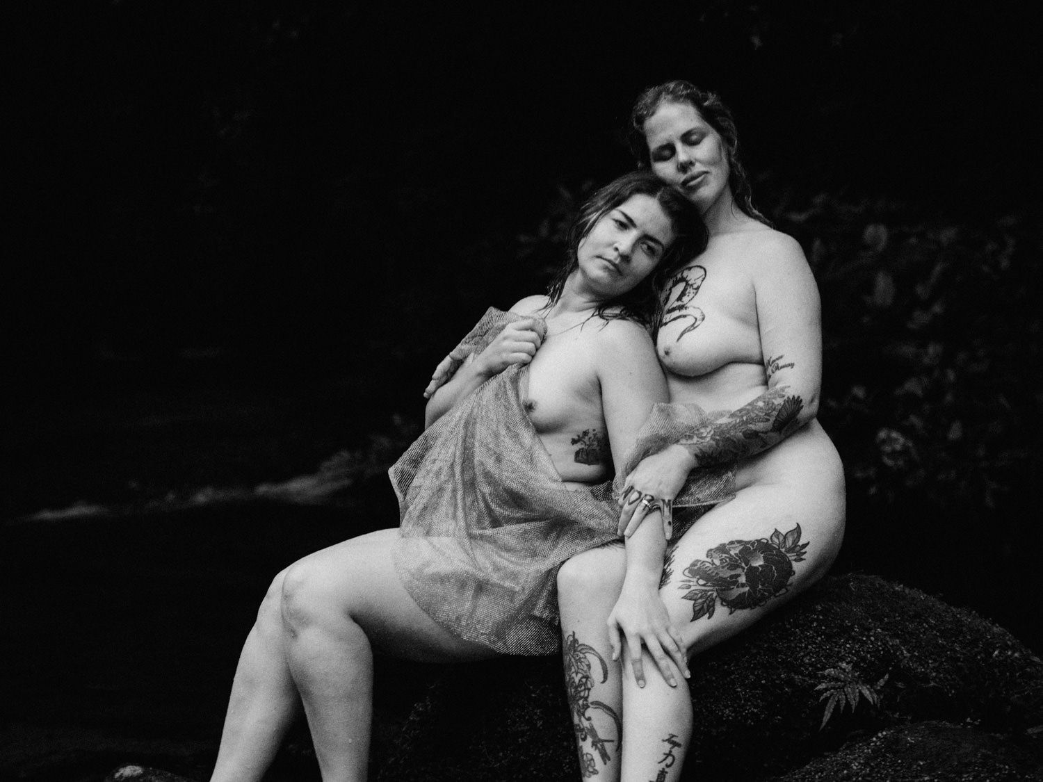 Two women with tattoos pose together on rocks near a stream in an artistic black and white nature photograph.