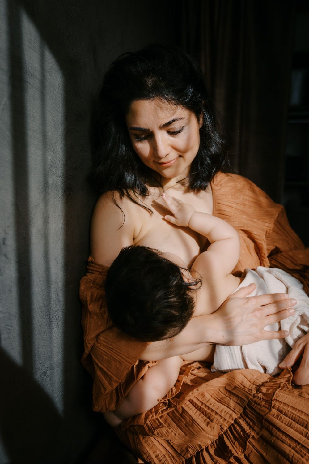 A mother tenderly breastfeeds her infant in warm natural light, wearing a rust-colored dress.