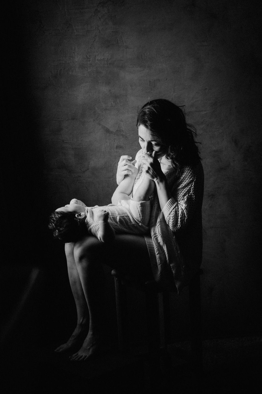 Black and white photo of a mother tenderly kissing her newborn baby while seated in a dimly lit studio.