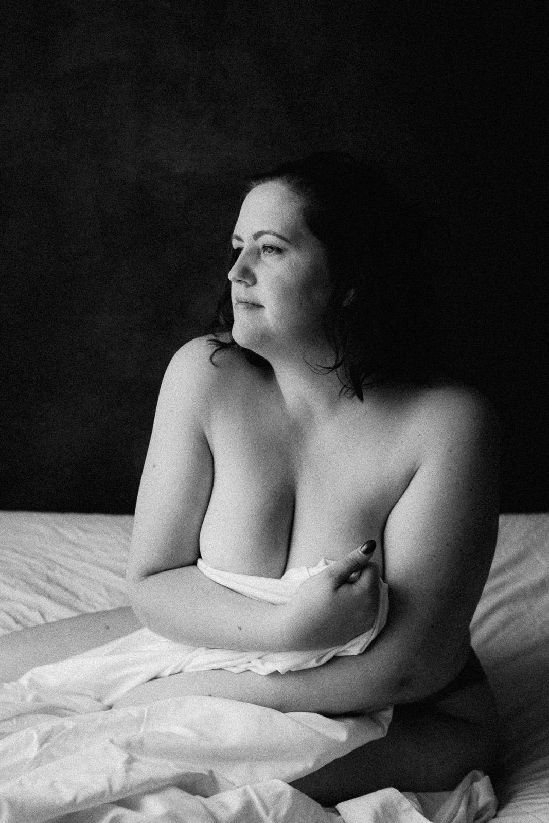 Black and white boudoir photo of a woman sitting on a bed, wrapped in white fabric, gazing away.
