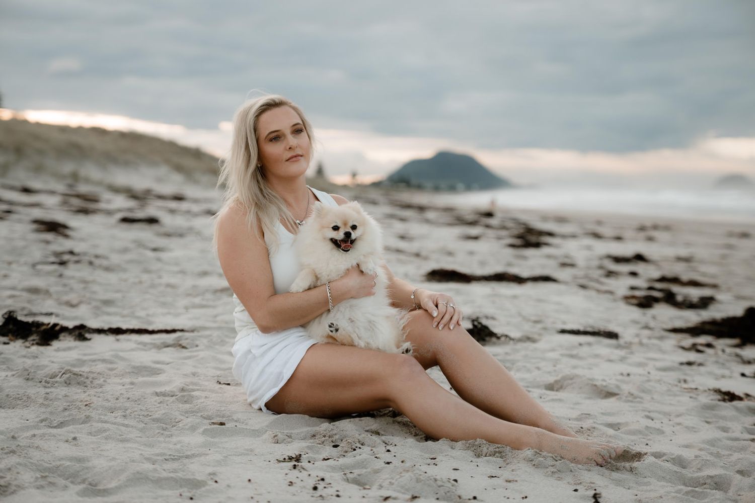 Blonde woman in white dress sitting on beach sand holding a fluffy cream Pomeranian dog with cloudy sky.